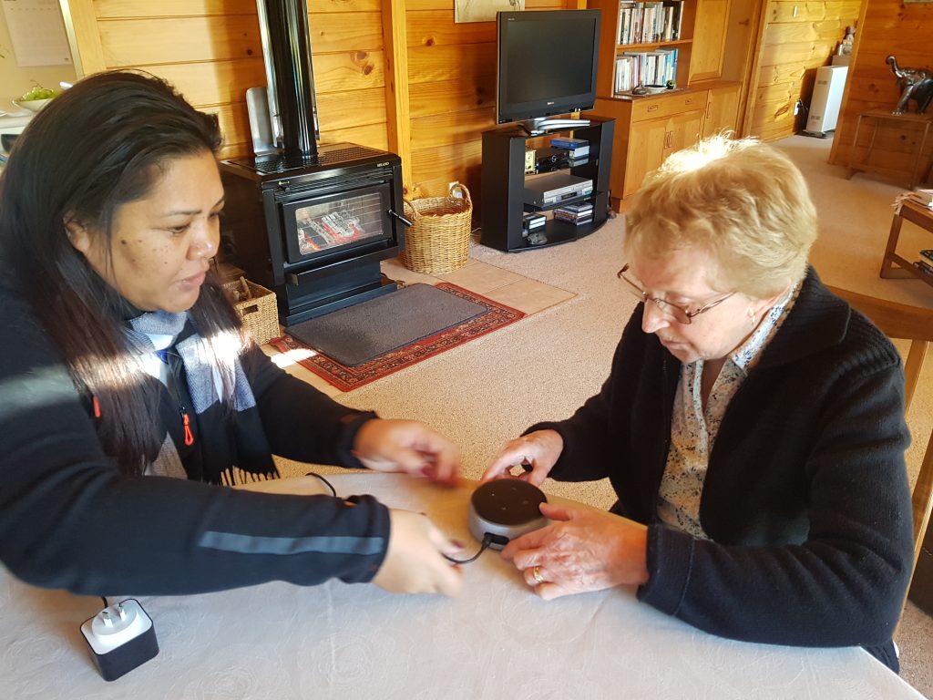 Blind Low Vision NZ. Two people, one elderly, sit at a table looking at an Amazon Echo dot.