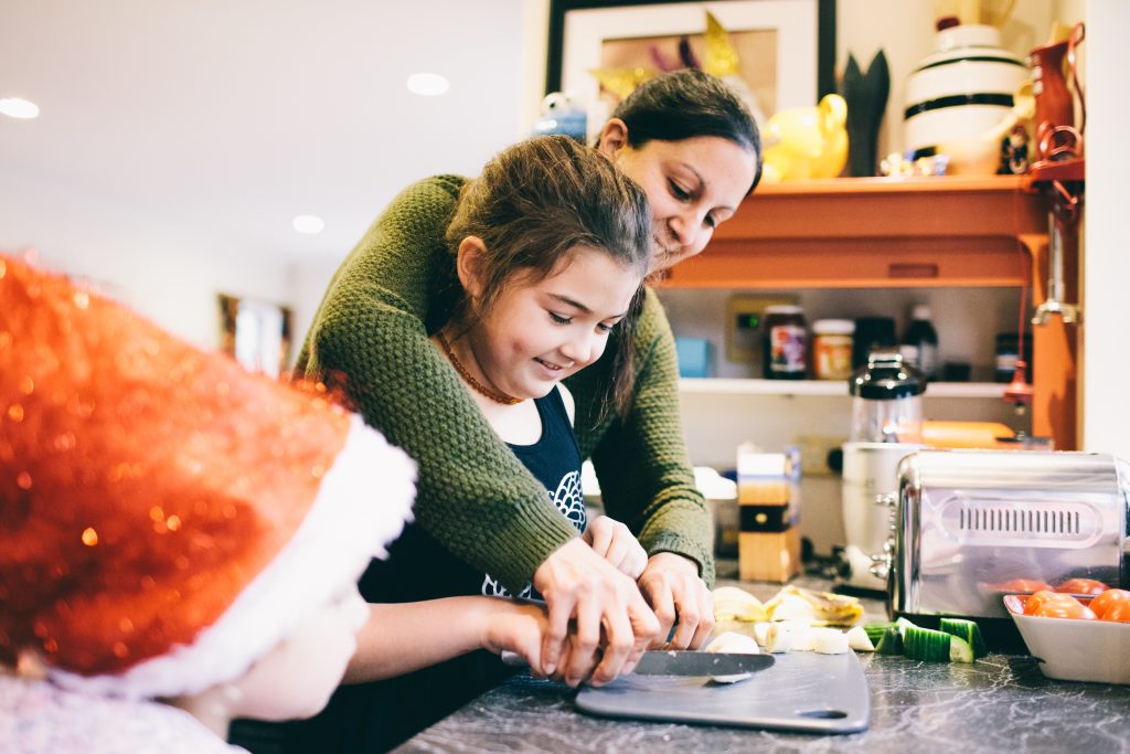 Blind Low Vision NZ. A mother and daughter in the kitchen chopping. A young child looks on.